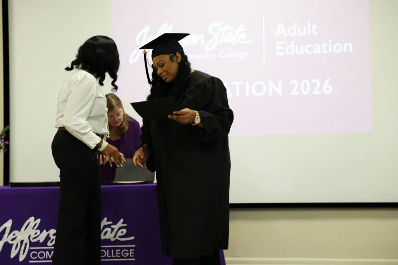 A graduate in a black cap and gown receives a certificate on stage from a woman during a Jefferson State Community College Adult Education ceremony.