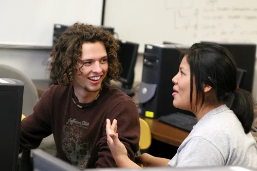 Two students sit at computer desks in a classroom, engaged in conversation. The student on the left is smiling with curly hair, while the student on the right gestures with her hand as she speaks.