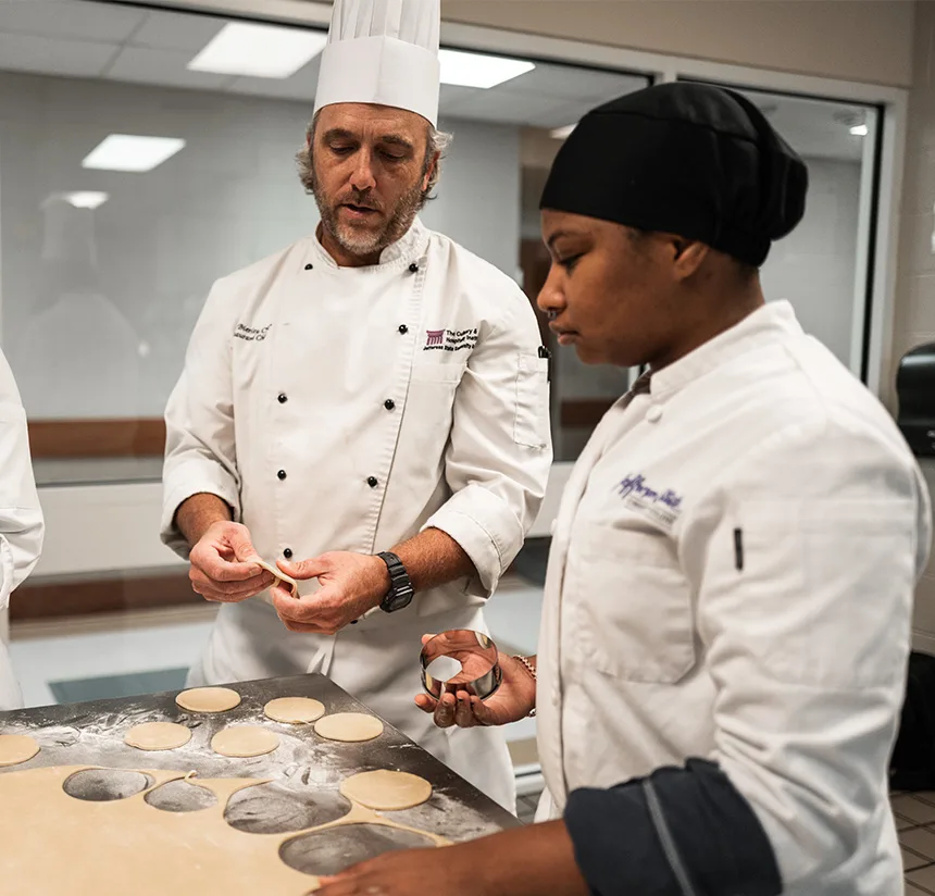A chef in a white uniform and tall hat demonstrates making dough circles to a student in a white jacket and black headscarf, in a professional kitchen setting with a flour-dusted table.