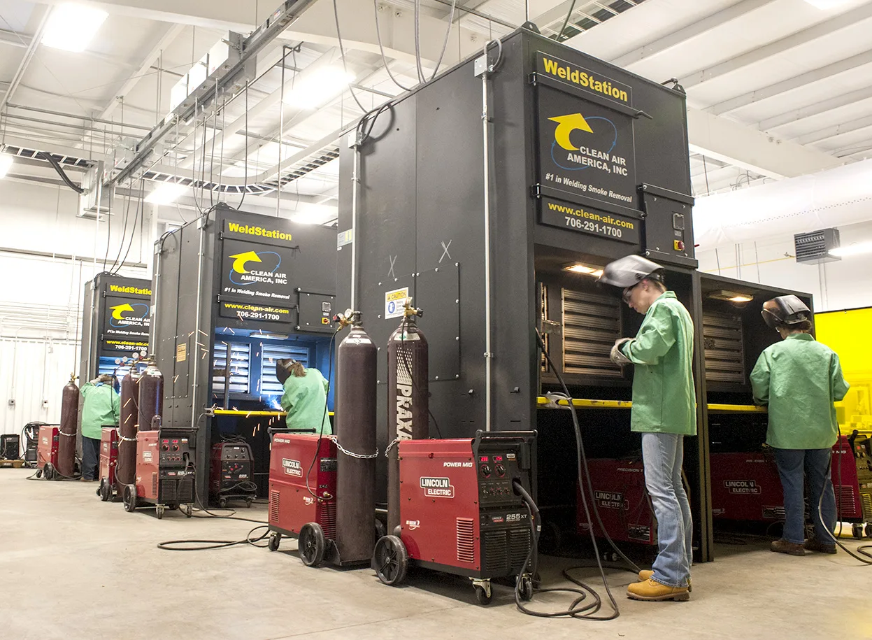 Three people in protective gear work at individual welding stations with large ventilation units and welding machines in an industrial workshop. The area is well-lit and organized, with safety equipment visible.