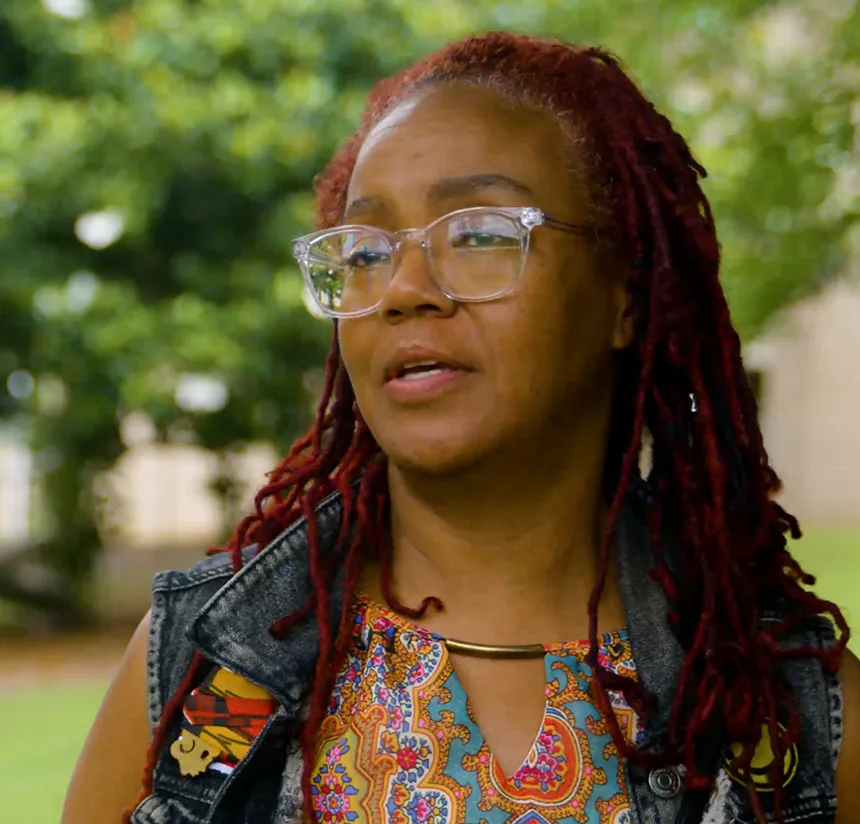 A person with long red dreadlocks, wearing glasses, a colorful patterned shirt, and a denim vest, stands outdoors with greenery in the background.