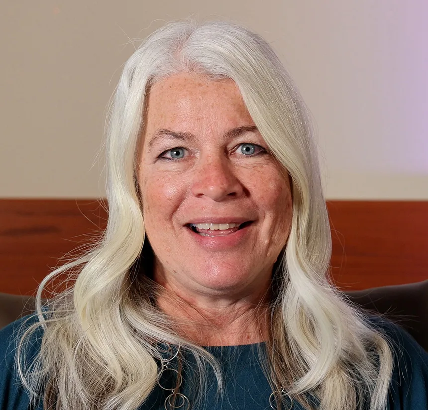 A smiling older woman with long white hair and blue eyes is wearing a dark green top and a necklace. She is indoors, sitting in front of a neutral-colored wall with wooden accents.
