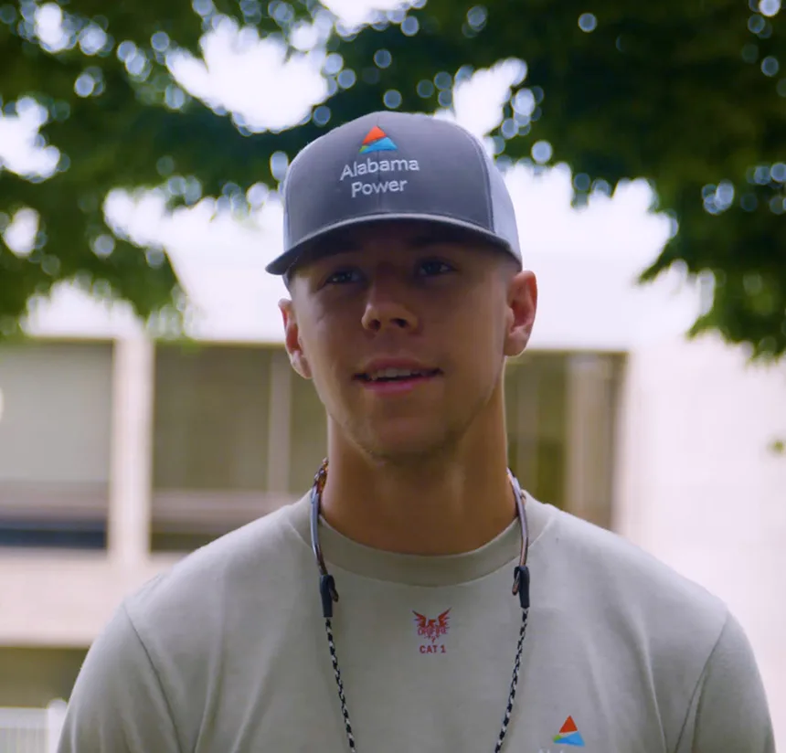 A young man wearing a gray Alabama Power cap, light gray shirt, and safety glasses around his neck stands outdoors with trees and a building in the background.