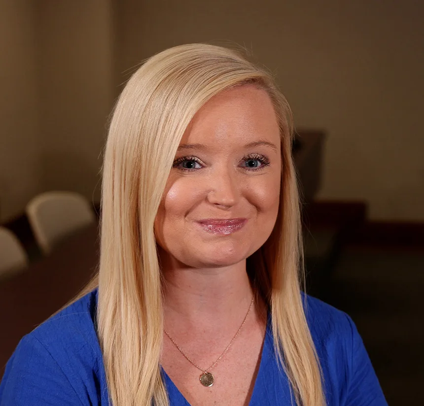 A woman with long straight blonde hair, wearing a blue top and a gold necklace, smiles at the camera while seated indoors. The background is softly blurred, showing chairs and a neutral-toned wall.