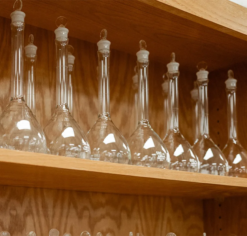Several clear glass volumetric flasks with stoppers are neatly arranged on wooden shelves against a wood-paneled background in a laboratory setting.