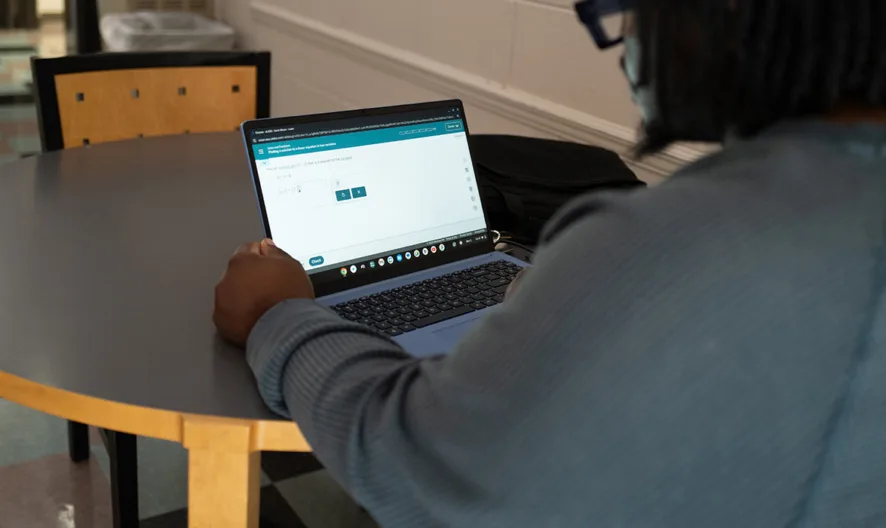 A person with shoulder-length hair sits at a round table, using a laptop in a hallway with checkered tile flooring. The laptop screen displays a webpage with a form or survey.