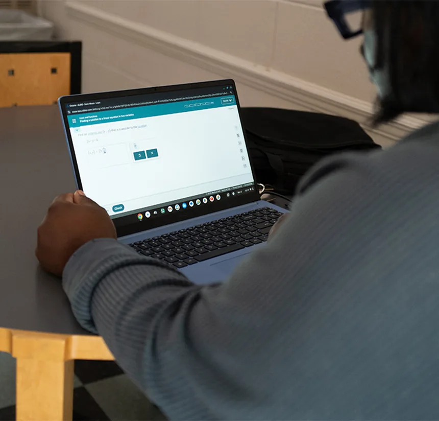A person sits at a table using a laptop, working on a math problem displayed on the screen. Their face is not visible, and they are wearing a long-sleeve shirt.
