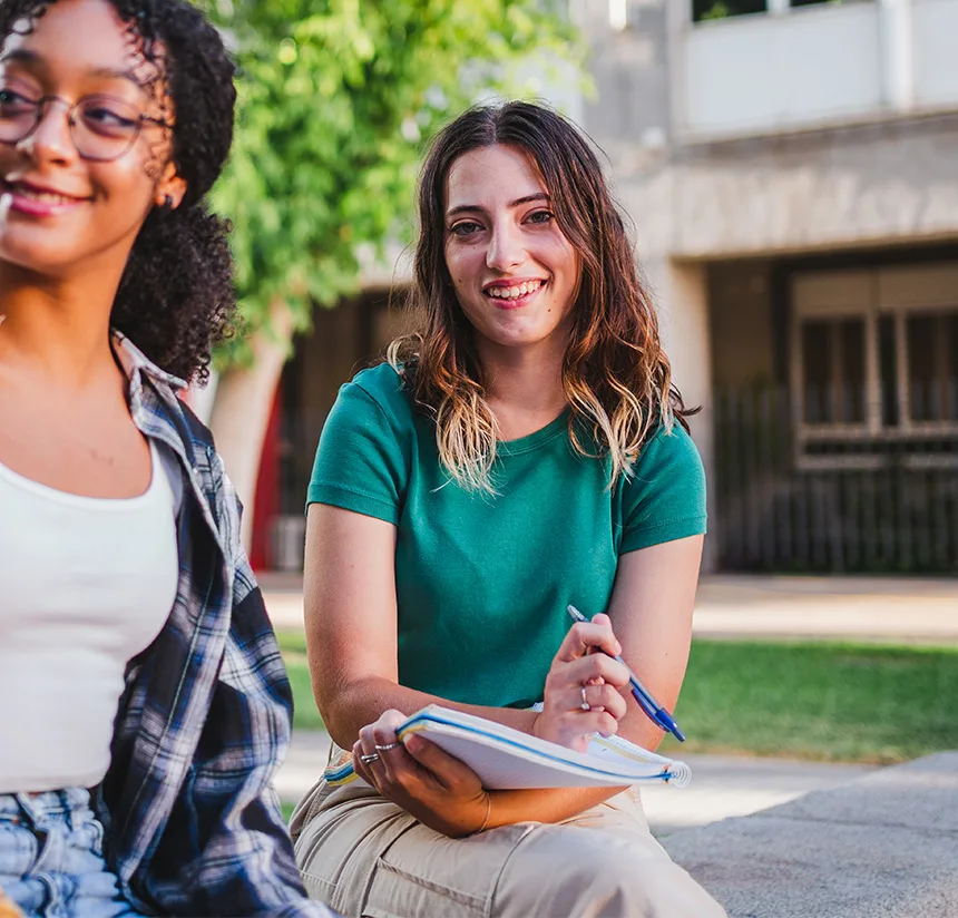 Two young women sit outdoors. One, wearing a green shirt, smiles at the camera while holding a notebook and pen. The other, in glasses and a plaid shirt, is partially visible. Green trees and a building are in the background.