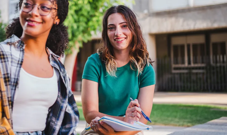 Two young women sit outdoors on a bench, smiling. One holds a notebook and pen, while both appear relaxed and happy, possibly studying together on a school or college campus with trees and buildings in the background.