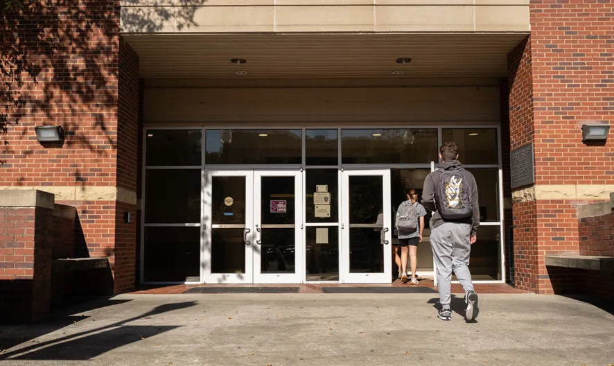 A student wearing a backpack walks toward the entrance of a brick building with glass doors on a sunny day, while another person stands near the doors.