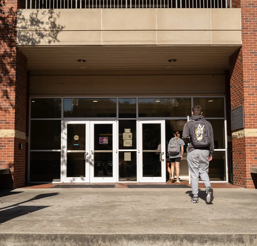 Two students with backpacks walk toward the entrance of a brick building with large glass doors on a sunny day.