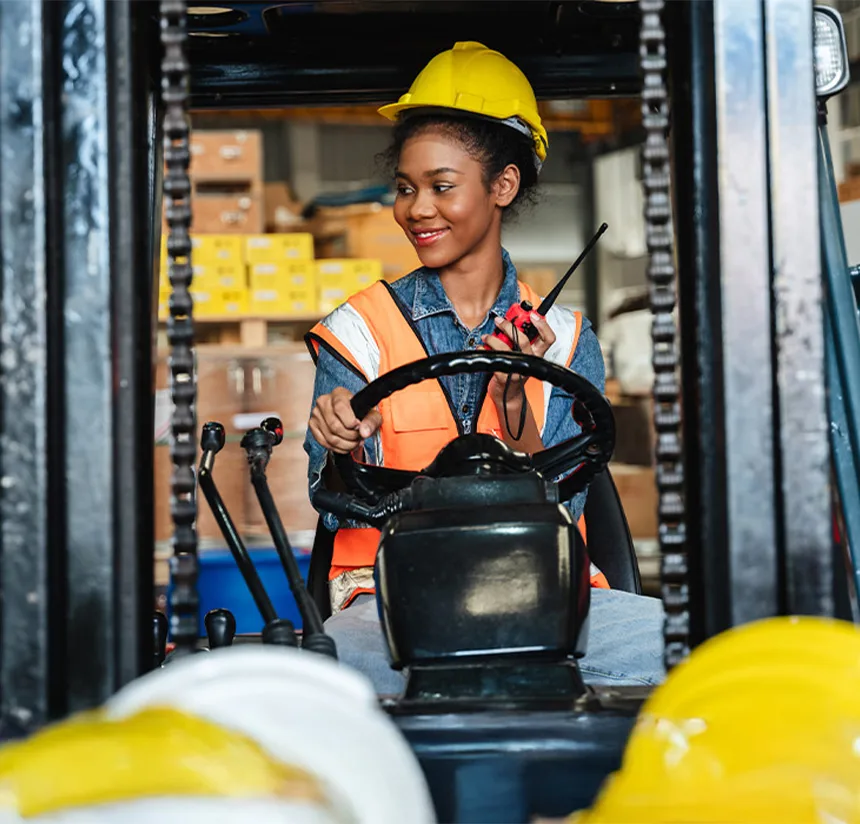 A woman wearing a yellow hard hat and orange safety vest sits at the controls of a forklift in a warehouse, smiling and holding a walkie-talkie. Boxes are stacked in the background.