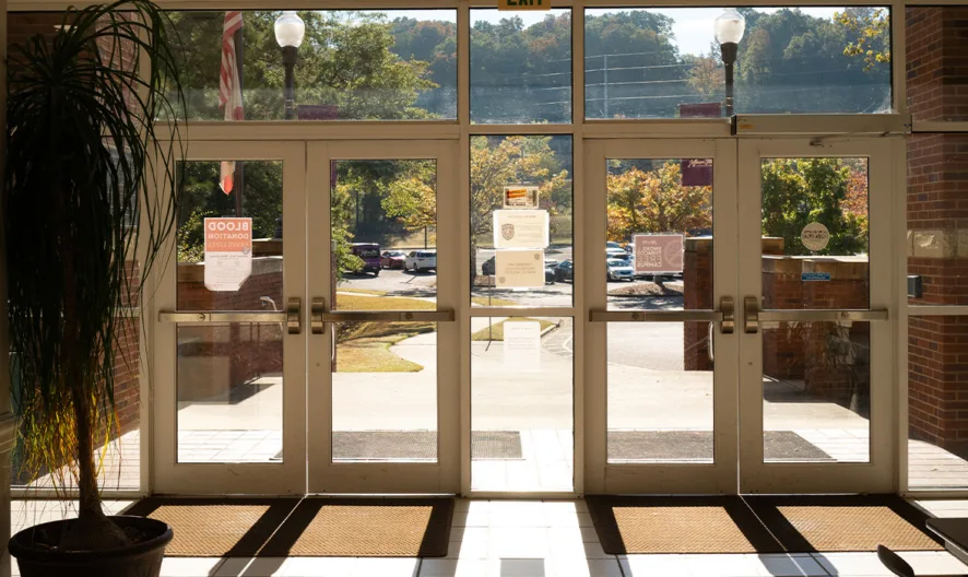 Sunlight streams through large glass doors at a building entrance, casting shadows on the floor. Outside, a parking lot and trees with fall foliage are visible. Bulletin boards and signs hang on the interior walls.