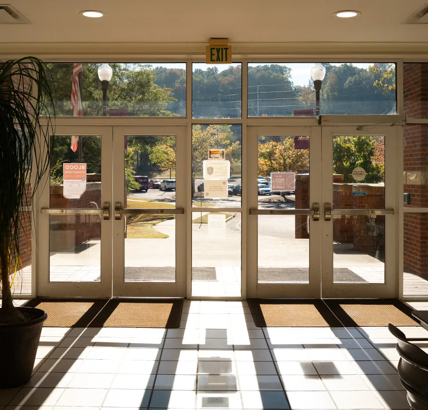 Sunlight streams through large glass double doors at a building entrance, casting shadows on the tiled floor. Outside, there are trees, cars, and a parking lot visible through the doors.