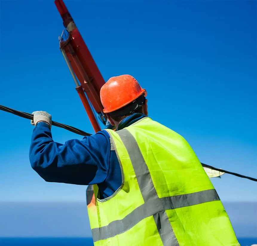 A construction worker in a fluorescent yellow safety vest and orange hard hat holds a cable while looking up at machinery against a clear blue sky.