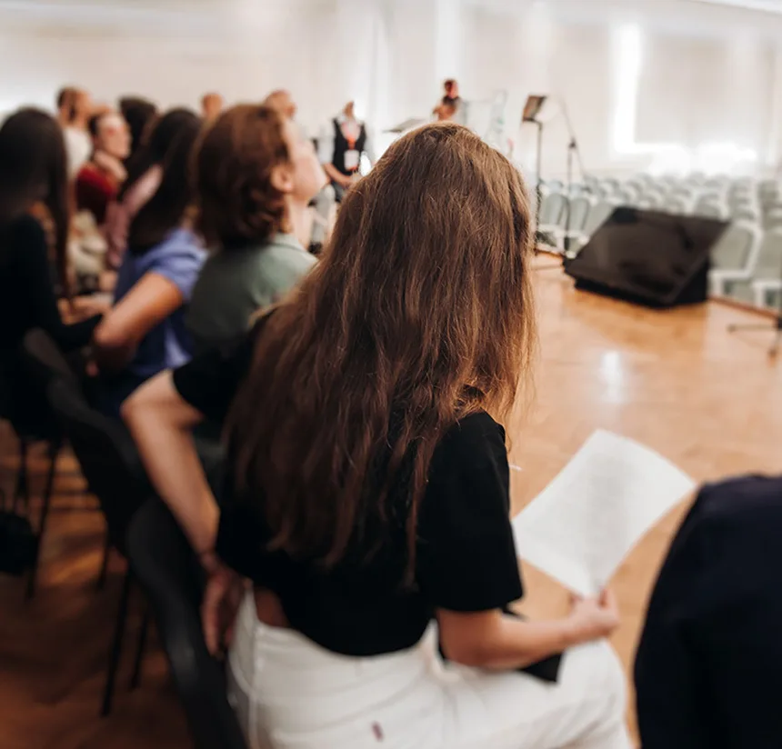A woman with long brown hair sits among others in rows of chairs, holding a sheet of paper, in a bright room with a stage and empty chairs in the background, suggesting an event or rehearsal.