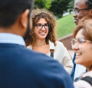 A woman with curly hair and glasses smiles while talking with a group of people outdoors. Trees and grass are visible in the background.