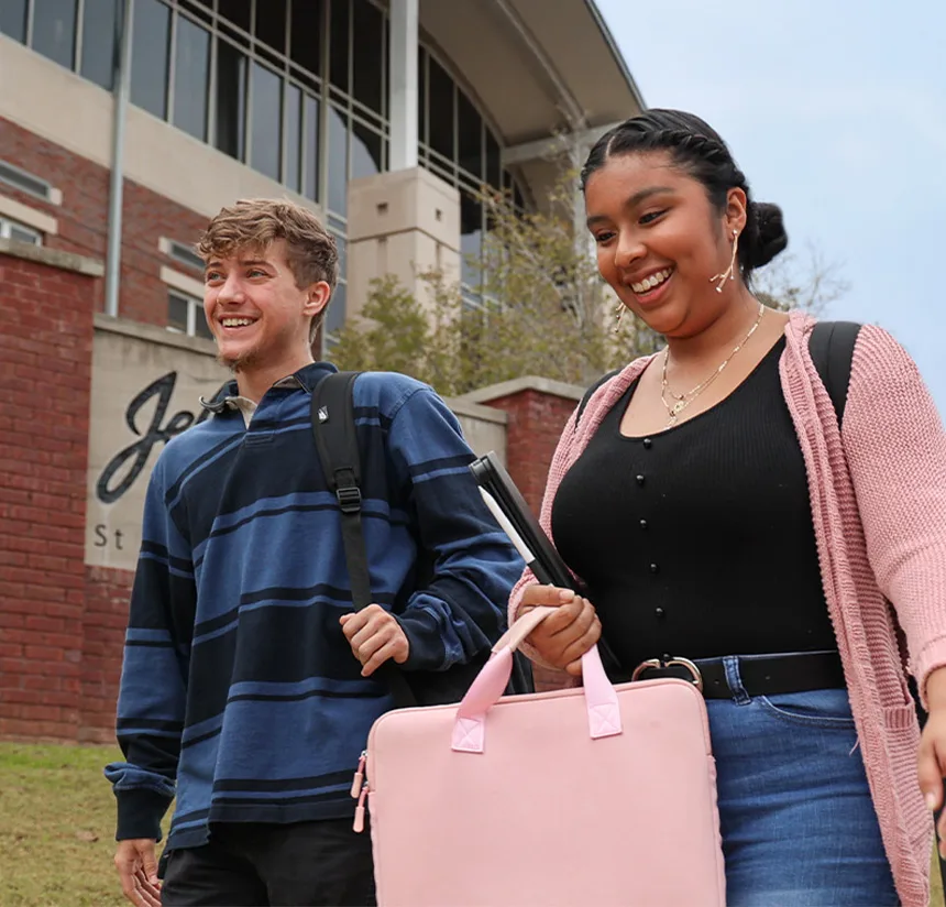 Two students, one male and one female, smiling and walking outside a red-brick campus building. The female carries a pink bag and notebook, while the male wears a striped sweater and has a backpack.