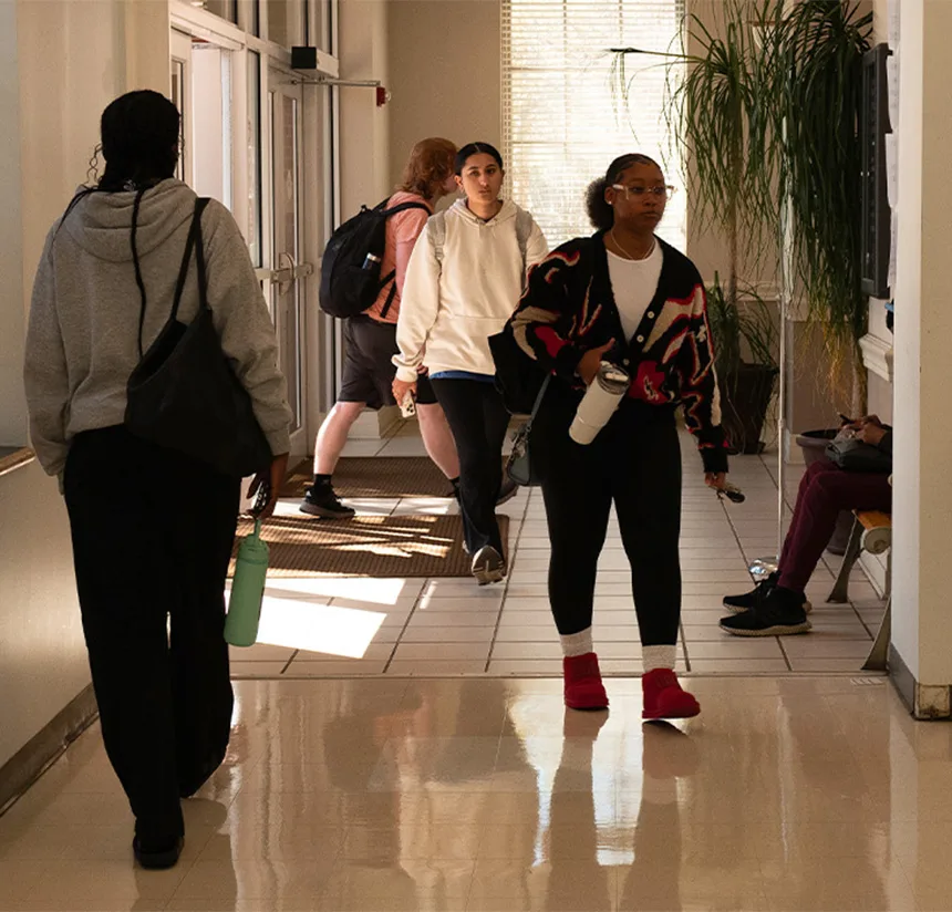 Several students walk through a sunlit hallway with large windows and potted plants. Some carry backpacks and water bottles, while another sits on a bench using a phone.