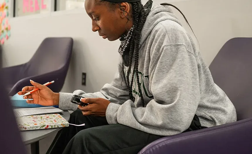 A person with braided hair, wearing a gray sweatshirt and black pants, sits in a purple chair writing in a notebook. They hold a device in one hand and appear to be studying in a brightly lit room.