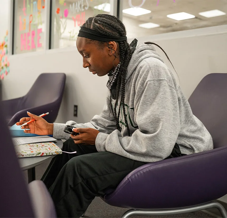 A person with braided hair, wearing a gray sweatshirt and black pants, sits in a purple chair writing in a notebook. They hold a device in one hand and appear to be studying in a brightly lit room.