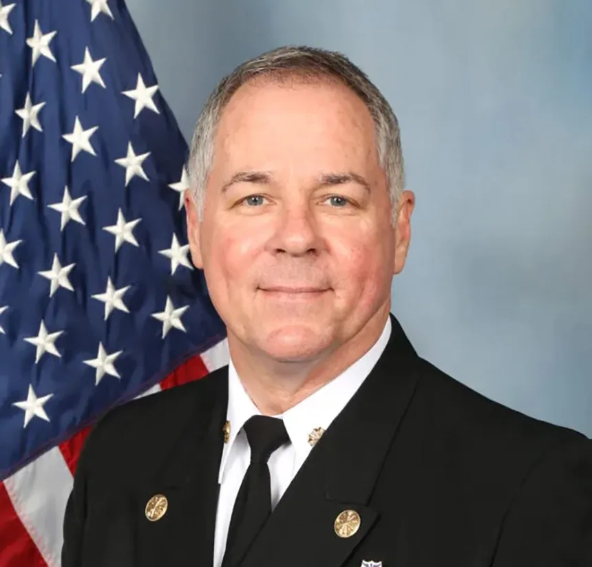 A man in a formal dark uniform with insignia poses in front of an American flag and a neutral background, smiling slightly.