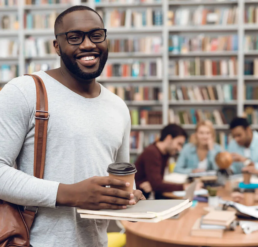 Smiling man with glasses, wearing a gray shirt and shoulder bag, holds coffee and folders in a library. In the background, people sit at a round table with bookshelves behind them.