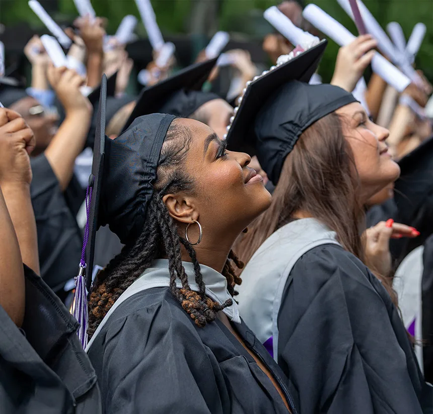A group of graduates in caps and gowns smiles and looks upward, holding rolled diplomas, during an outdoor graduation ceremony.