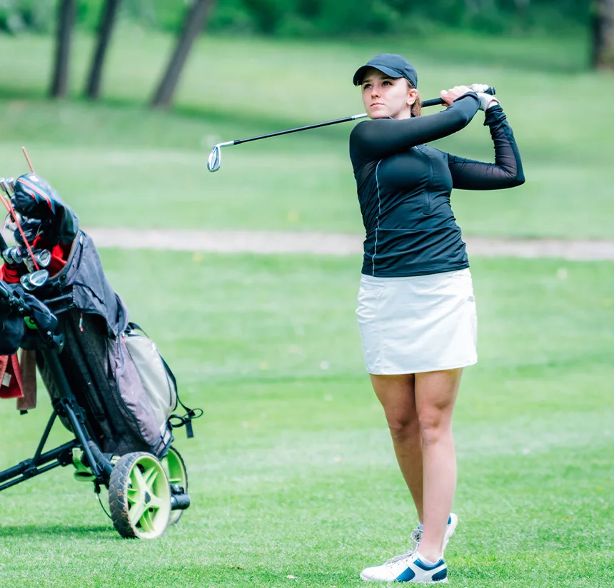 A woman wearing a black long-sleeve top, white skirt, and cap swings a golf club on a golf course; a golf bag on a push cart is beside her.