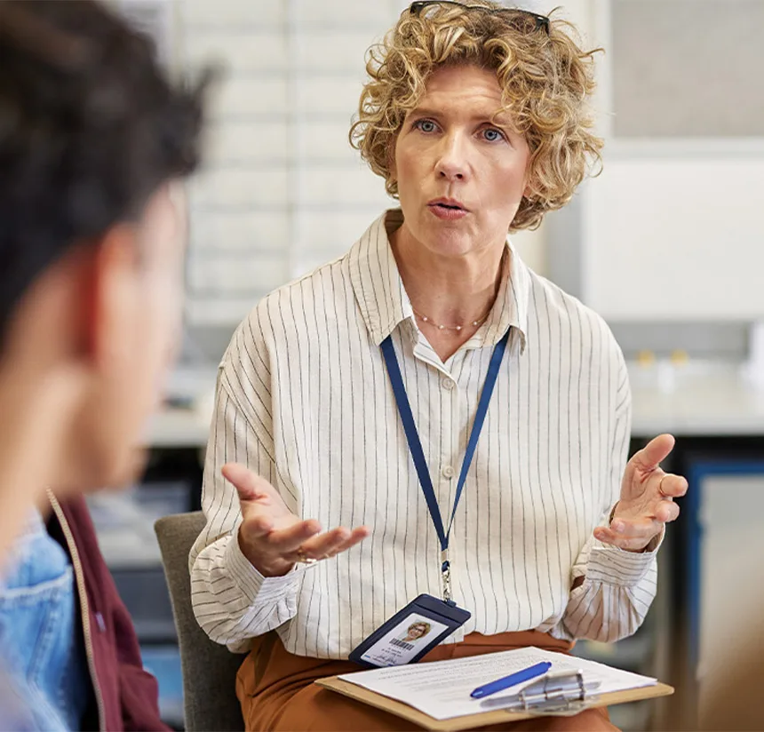 A woman with curly blonde hair wearing a striped shirt and lanyard sits indoors, holding a notebook and pen, and speaks with expressive hand gestures to a person facing her.
