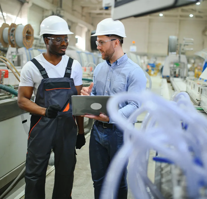 Two male factory workers wearing hard hats and safety glasses stand in a manufacturing facility. One holds a laptop and discusses something with the other, who wears overalls and gloves. Machinery and equipment are visible in the background.