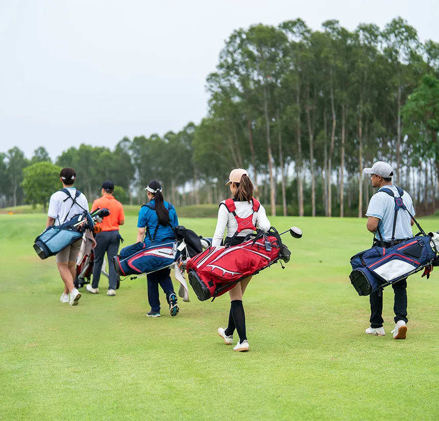 A group of five golfers, carrying golf bags, walk together on a green golf course bordered by trees under a cloudy sky.