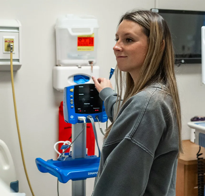 A woman in a gray sweater stands in a medical room, pointing at a blue medical monitor. There are various medical devices and supplies on the wall behind her.