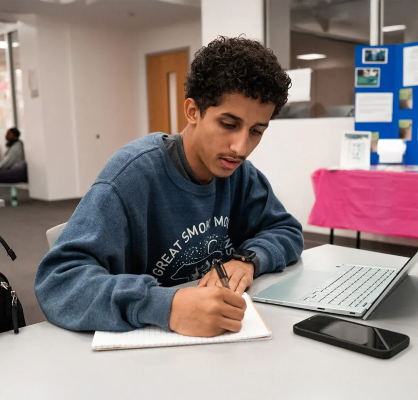 A young man sits at a table, writing in a notebook with a pen. He has a laptop and smartphone on the table and is wearing a blue sweatshirt. A colorful display board is visible in the background.