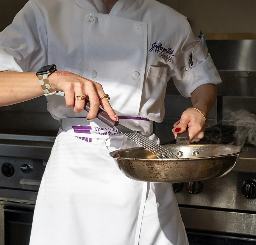 A chef in a white uniform uses a whisk to stir food in a steaming pan over a stove, wearing a smartwatch and rings, with sunlight highlighting their hands and utensils.