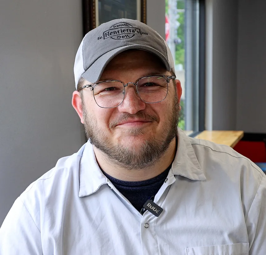 A smiling man with a beard and glasses wears a gray cap, light gray shirt, and a microphone clipped to his collar, sitting indoors by a window and wooden table.