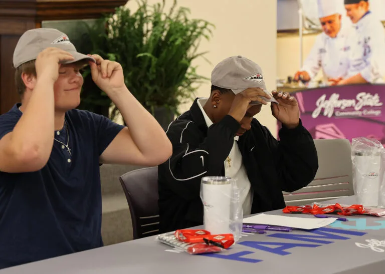 Two young people sit at a table, each putting on a gray hat. The table has papers, cups, and red items on it. In the background, there is a plant and a poster showing chefs at Jefferson State Community College.