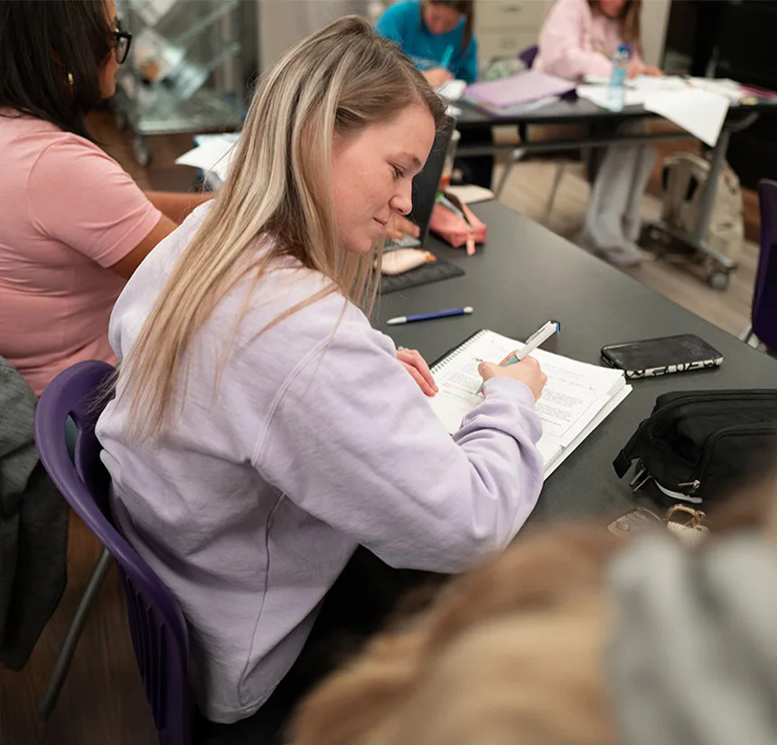 A young woman with long blonde hair sits at a desk in a classroom, writing in a notebook. She wears a light purple sweatshirt. Other students and study materials are visible in the background.