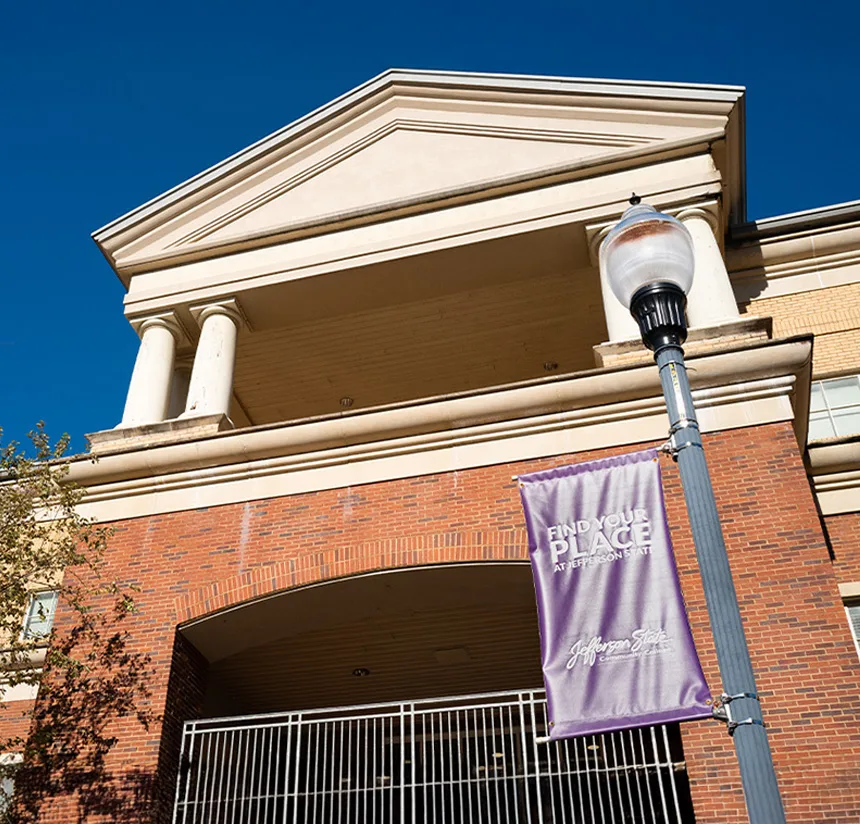 A red brick building with white columns and a triangular pediment under a clear blue sky. In front, a streetlamp holds a purple banner that reads, Find Your Place at Jefferson State.