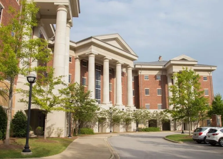 A large brick building with tall white columns and multiple windows, surrounded by trees and a few parked cars, under a partly cloudy sky.