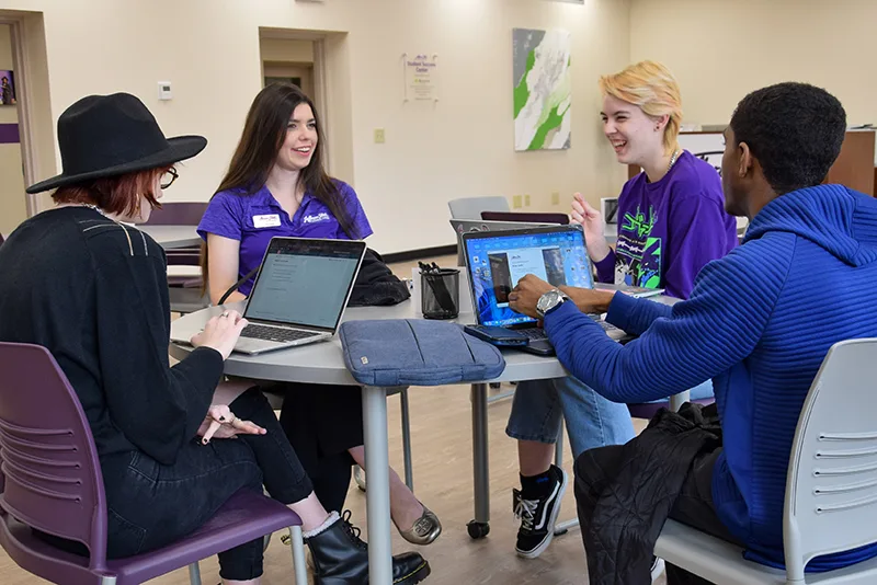 Four students sit around a table, working on laptops and talking. Two wear purple shirts, one wears a blue sweater, and one wears black with a hat. The setting is a bright, modern classroom or study space.