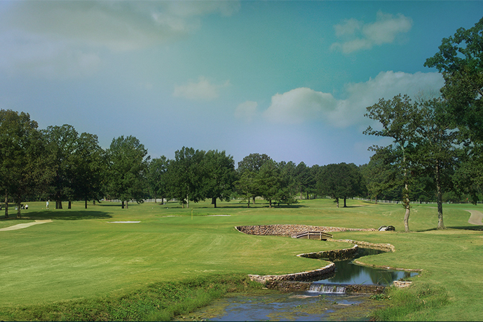 A scenic view of a golf course with lush green fairways, scattered trees, sand bunkers, and a small winding creek with a stone bridge under a partly cloudy blue sky.