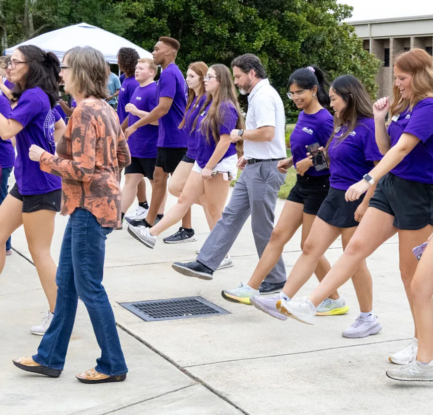 A group of people, mostly young women in matching purple shirts, march or dance in step outdoors, lifting their right legs, while two adults walk beside them. Trees and a building are visible in the background.