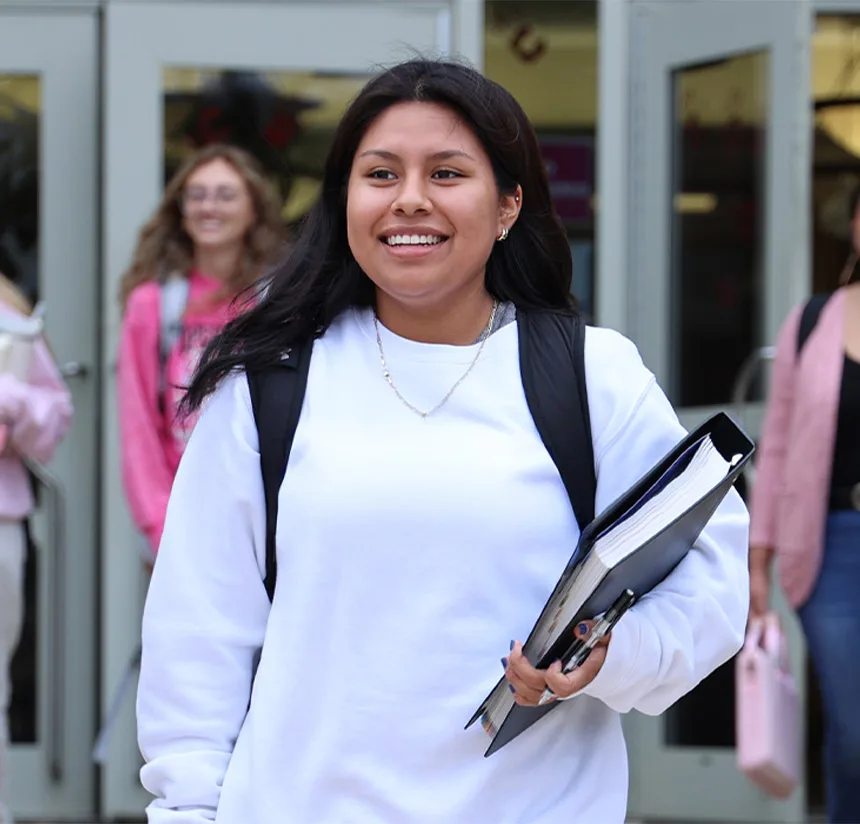 A young woman with long dark hair, wearing a white sweatshirt and backpack, smiles while holding a binder. She is walking outside with other students in the background near glass doors.