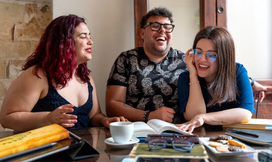 Three friends sit at a table, laughing and talking together. Open books, notebooks, coffee, and snacks are spread out in front of them, suggesting a casual study or work session in a cozy, well-lit room.