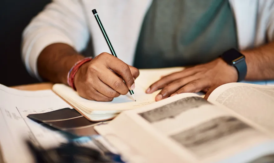 A person wearing a smartwatch writes in a notebook at a desk with an open book and papers spread out in front of them.