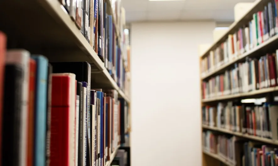 Bookshelves filled with colorful books line both sides of a narrow aisle in a library, with a bright, out-of-focus wall and ceiling lights visible at the far end.