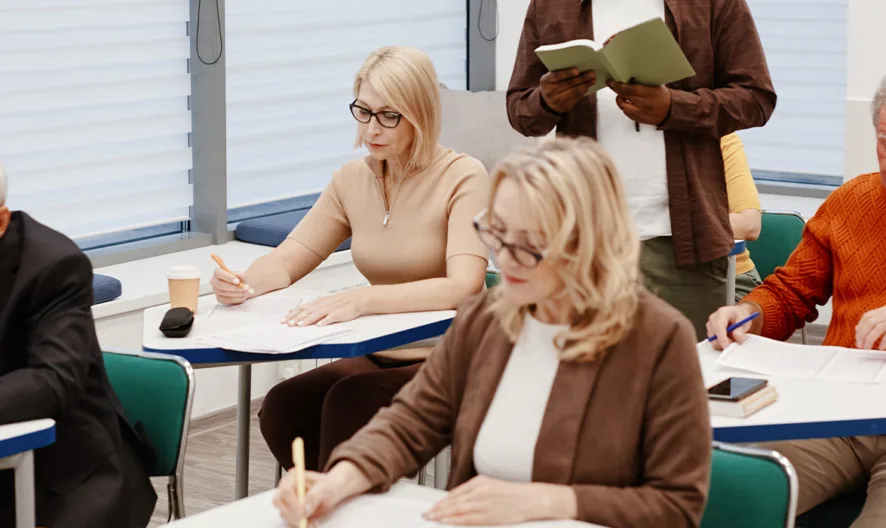 A group of older adults sit at desks in a classroom, writing on papers, while a standing person reads from a book at the front of the room.