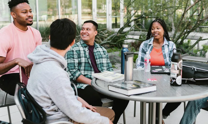 Five young adults sit around an outdoor table, talking and smiling. Books, notebooks, and water bottles are on the table. Greenery and glass buildings are visible in the background.