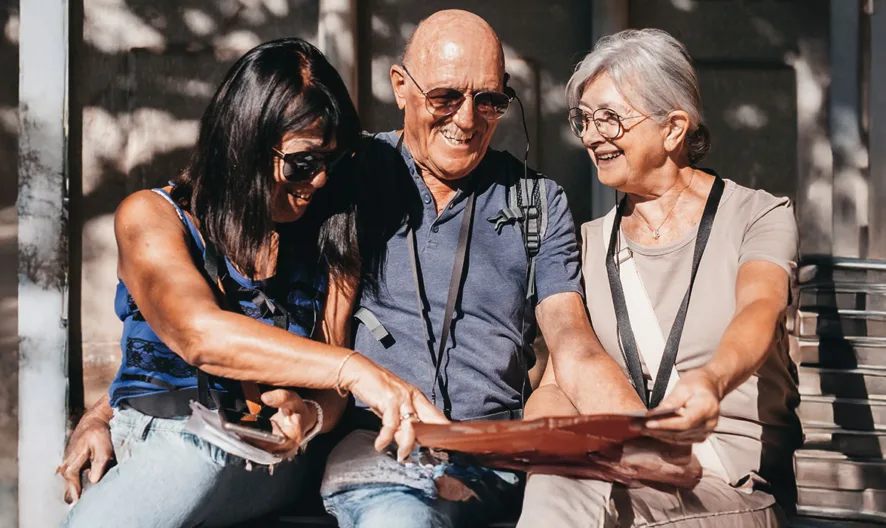 Three people sit on a bench outdoors, smiling and looking at a photo album together. The two women and one man appear happy and engaged, sharing memories on a sunny day.